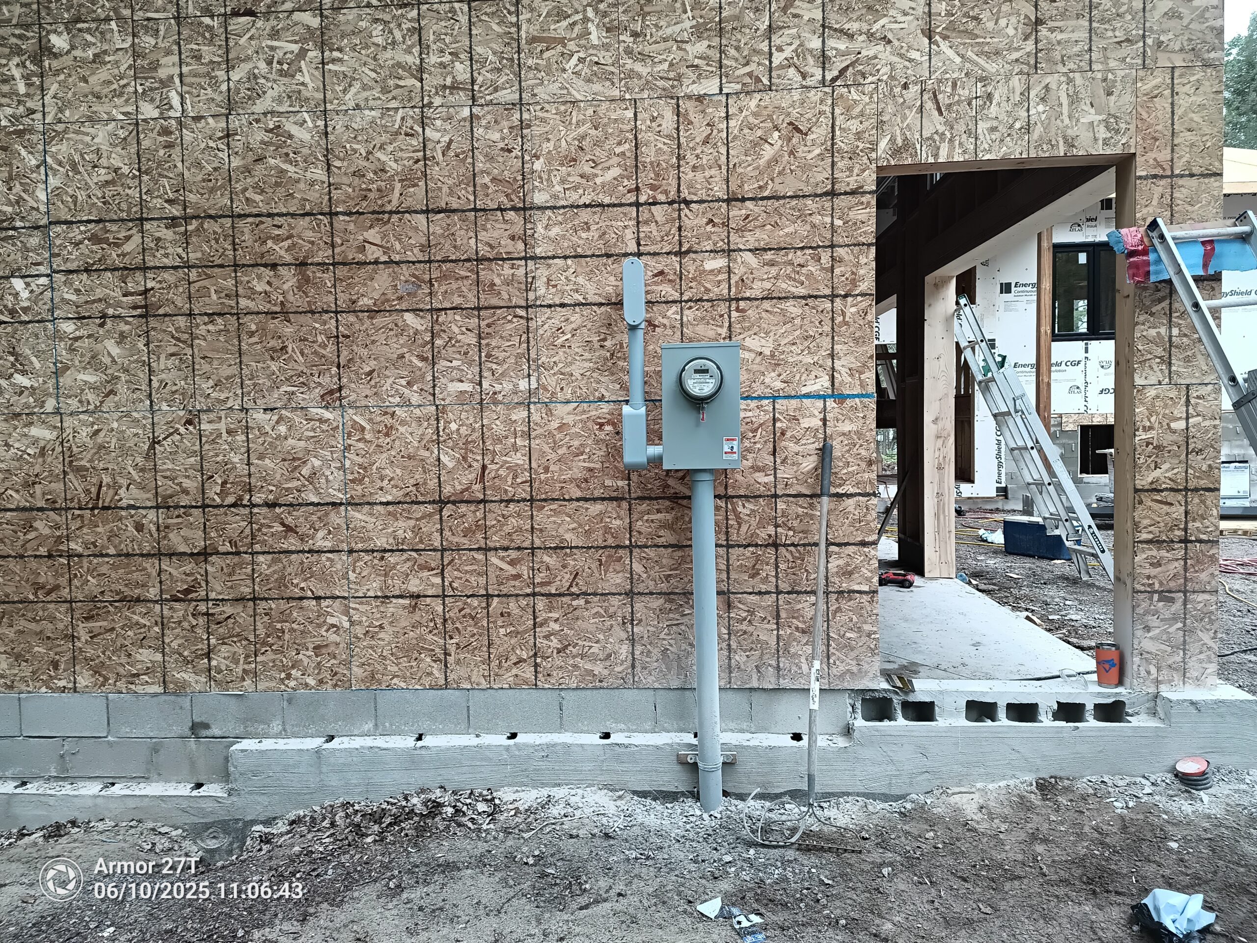Partially constructed house exterior with OSB sheathing, visible utility meter box and conduit, cinder block foundation, and doorway under construction with ladders inside.
