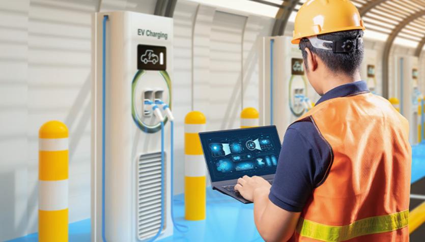 Engineer in safety vest and helmet using a laptop to monitor EV charging station performance.