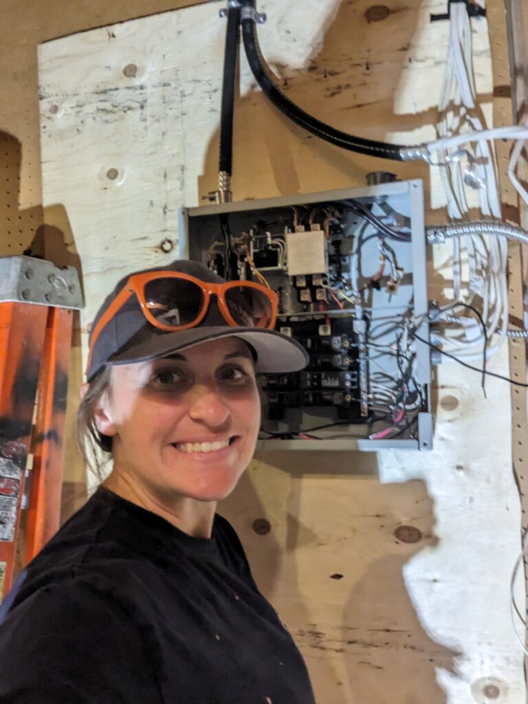 A smiling female electrician wearing a cap and sunglasses, standing in front of an open electrical panel with exposed wiring.