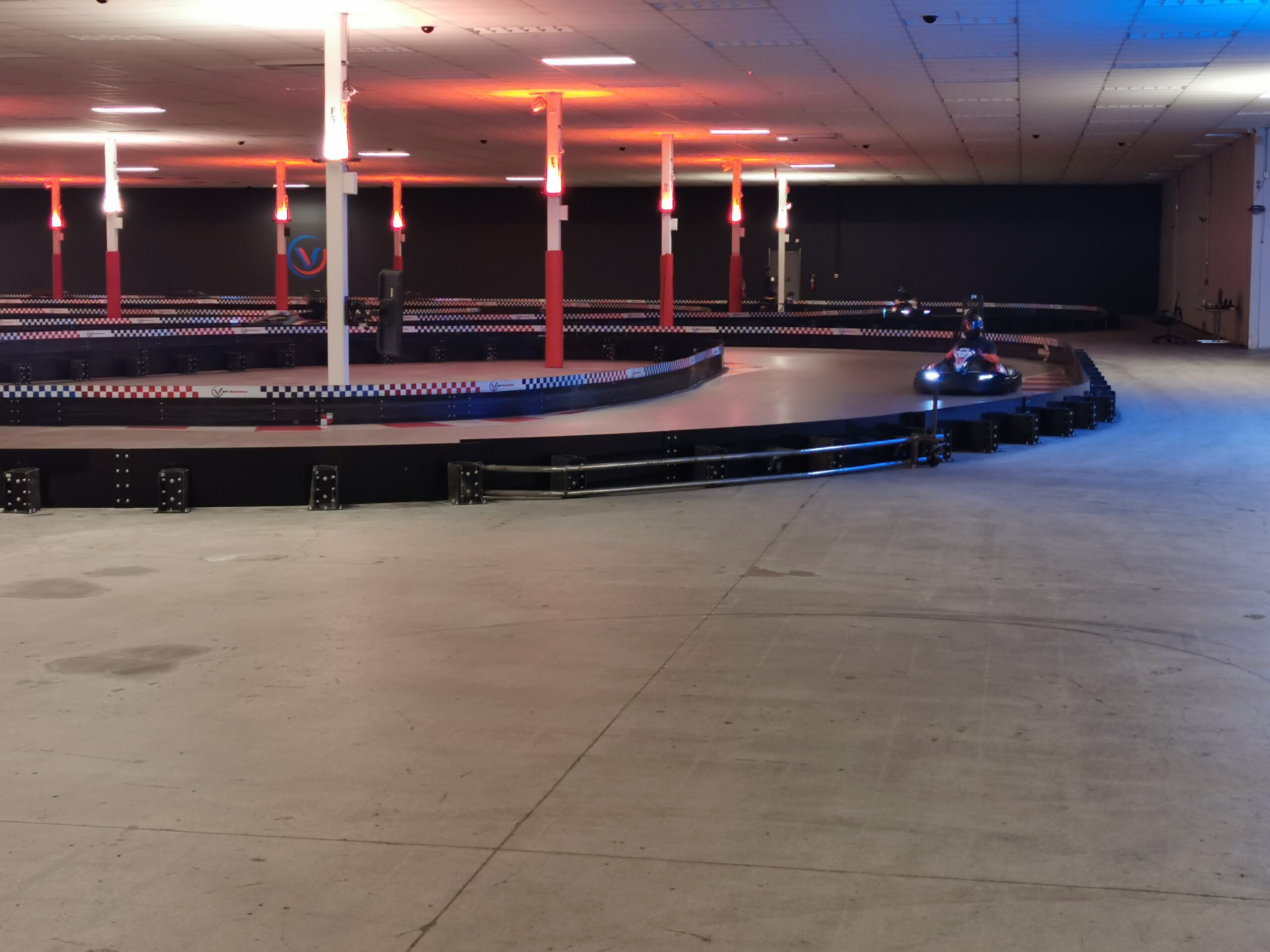 Indoor electric go-kart racing track with bright overhead lights, safety barriers, and a go-kart approaching a corner.