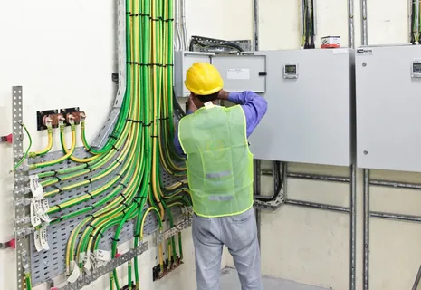 An electrician wearing a hard hat and safety vest works on a complex electrical panel with numerous wires and conduits.