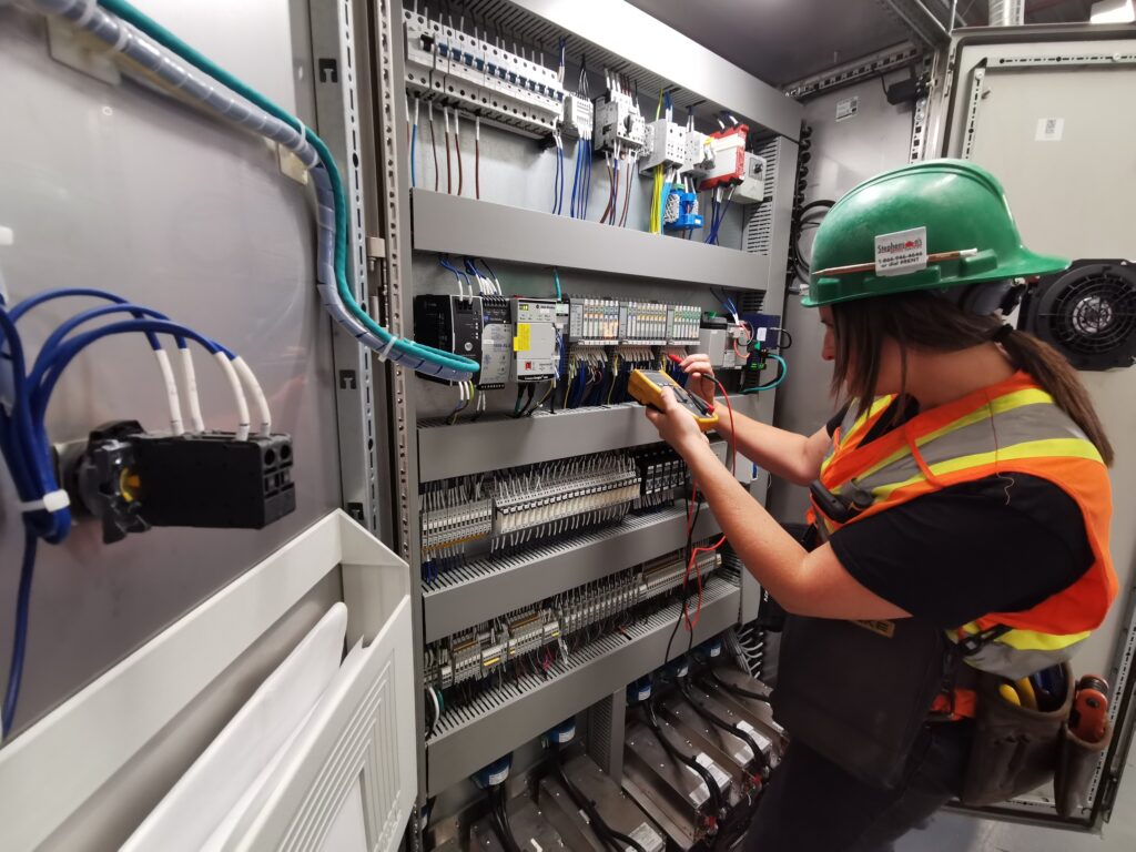 Female electrical technician wearing a safety helmet and vest testing control panel wiring with a multimeter inside an industrial electrical cabinet.