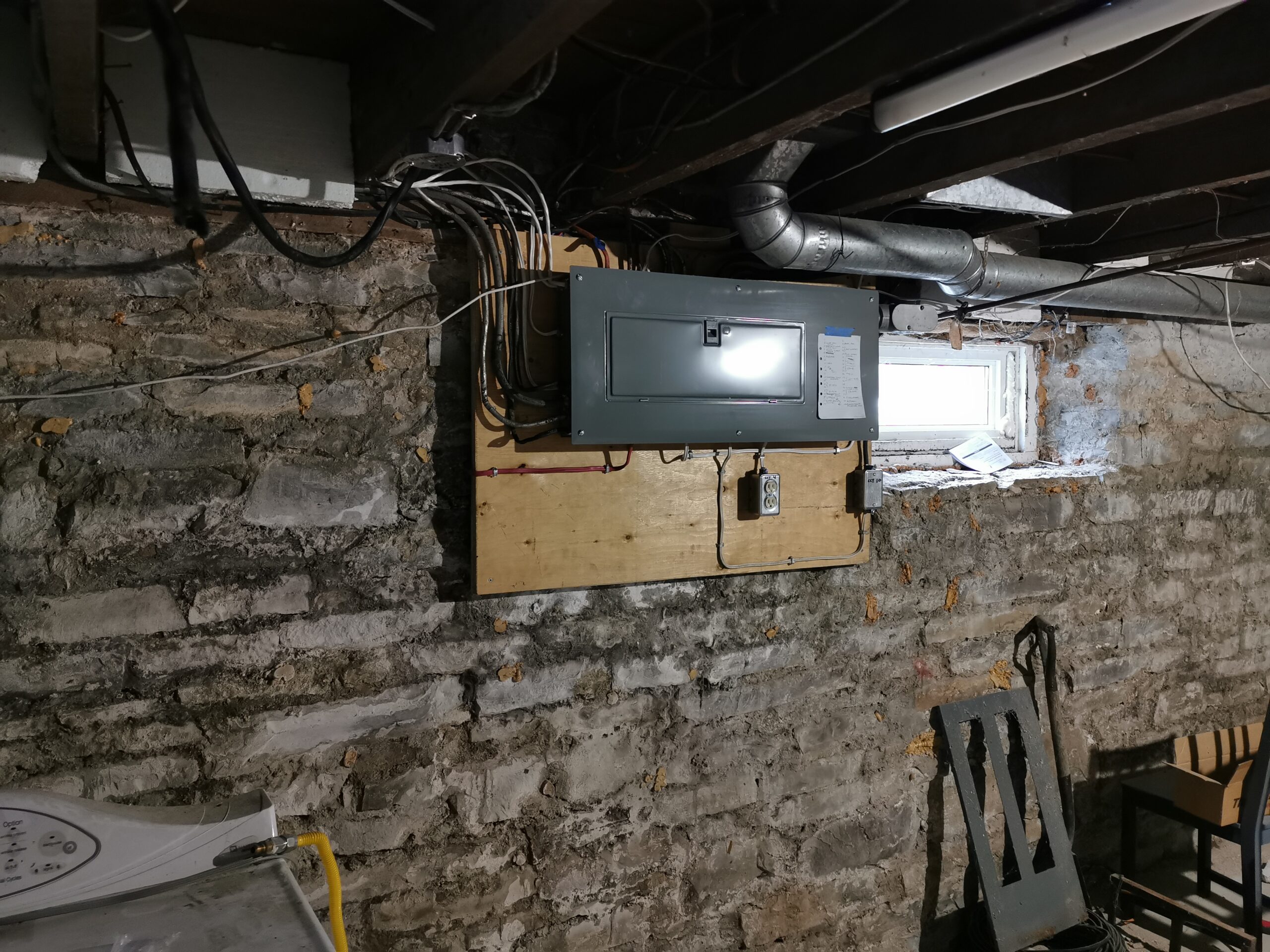 Exposed electrical panel installed on a wooden board in a basement with stone walls, visible wiring, ductwork, and natural light from a small window.