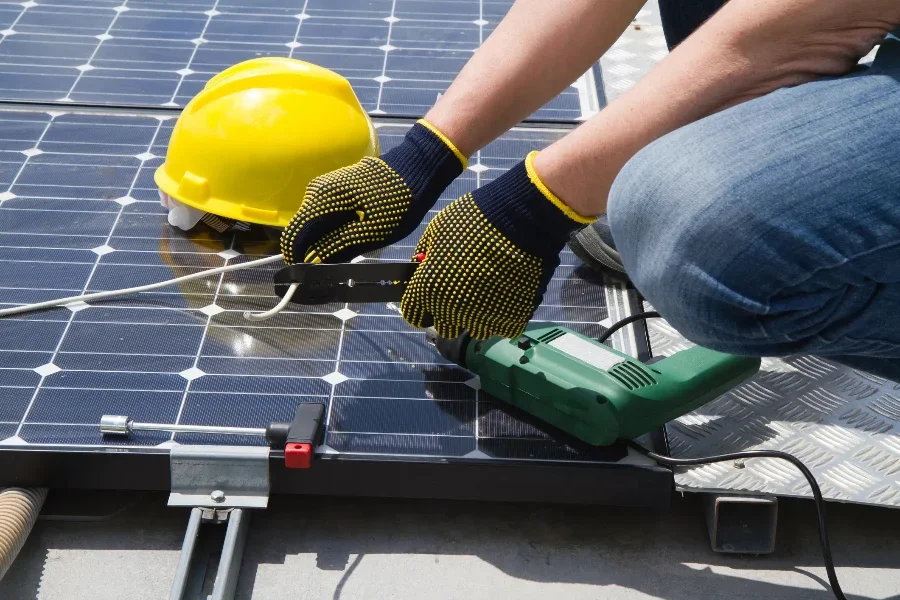 A close-up of a technician's gloved hands using a power tool to install a solar panel, with a yellow hard hat nearby.