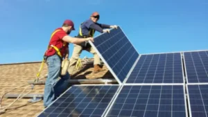 Two technicians, wearing safety harnesses and helmets, work together to install a solar panel on a residential rooftop. The sky is clear and blue, and other panels are already in place, forming a neat grid.