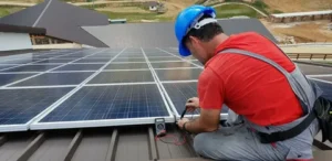 A technician wearing a blue hard hat and a red shirt kneels on a brown metal rooftop, using a multimeter to check the wiring of a solar panel. The solar panel array covers the roof, with a modern house and a hilly landscape visible in the background.