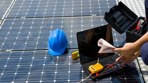 A close-up, high-angle shot of a person working on a solar panel array. They are using a multimeter and a laptop to test the electrical connections. A blue hard hat and a level are also on the panels.