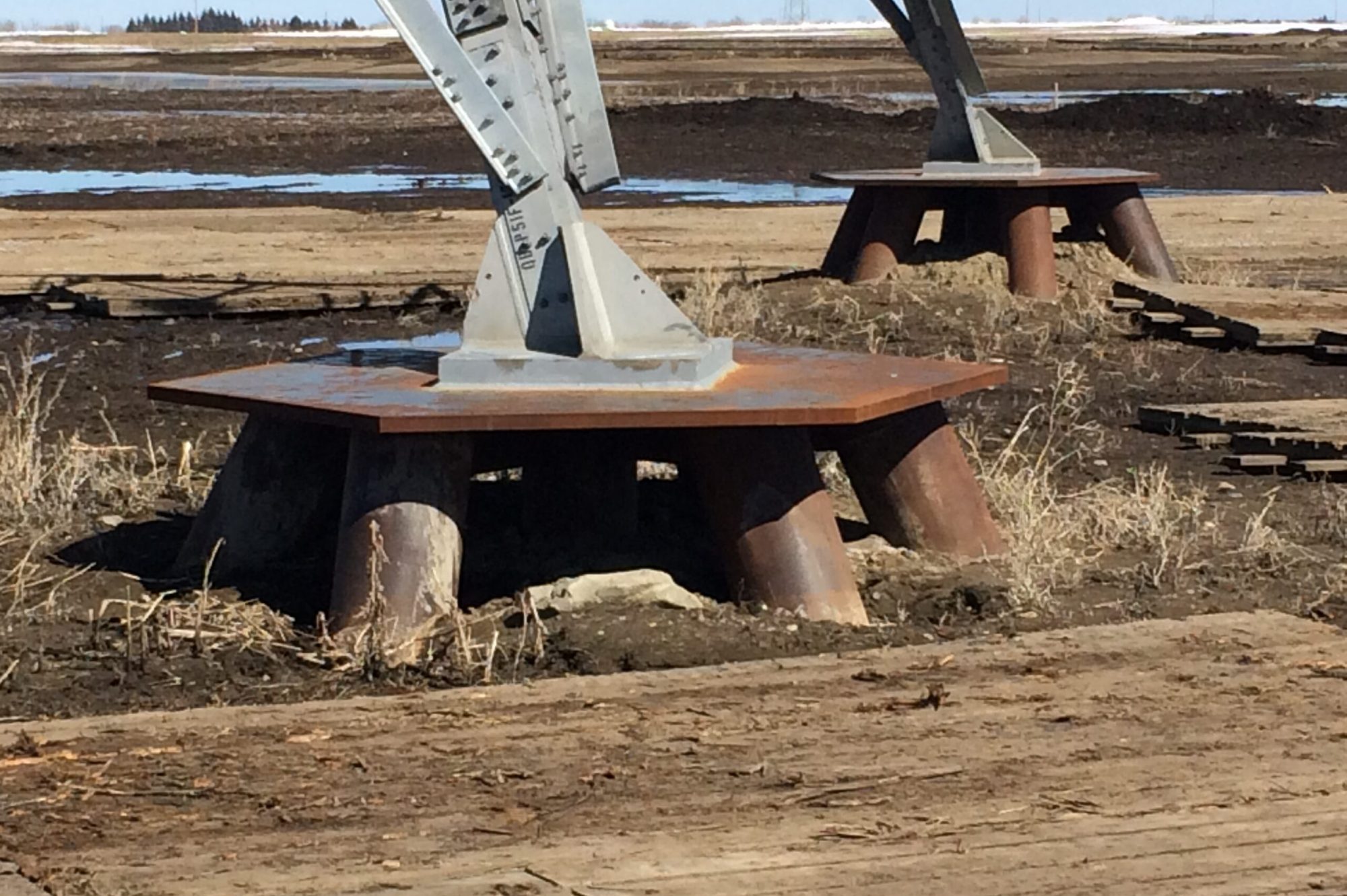 Transmission Tower Foundation with Steel Pile Supports Steel transmission tower foundation with angled concrete base plates mounted on large steel piles in a muddy construction site.