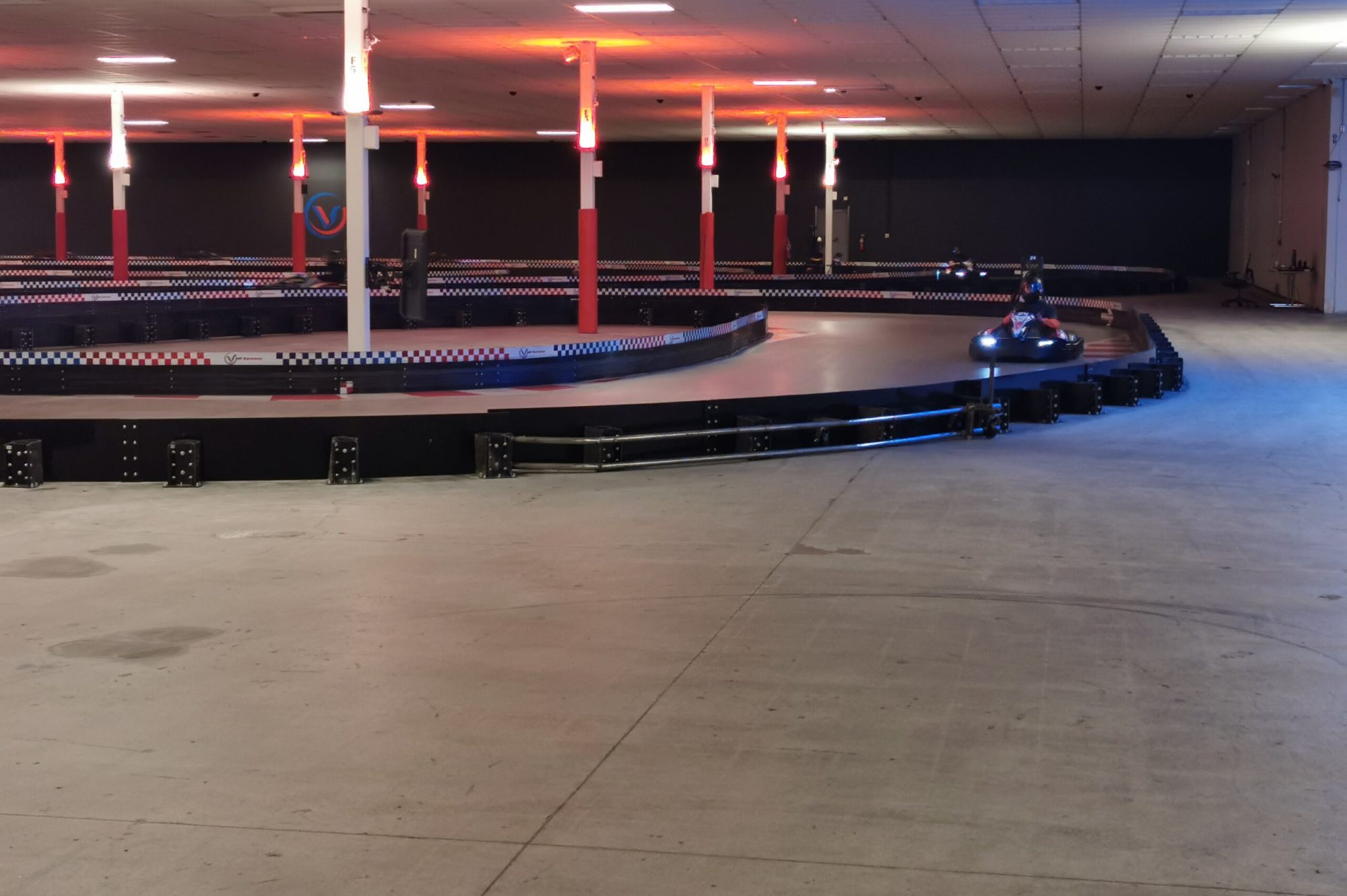 Indoor Electric Go-Kart Racing Track with Safety Barriers Indoor electric go-kart racing track with bright overhead lights, safety barriers, and a go-kart approaching a corner.