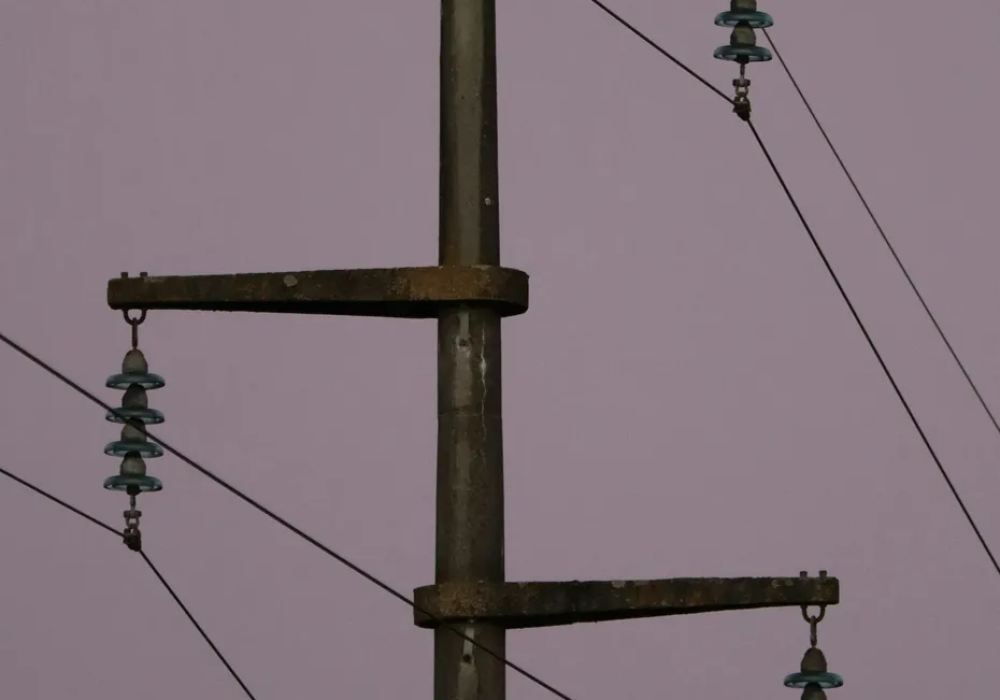 A close-up of a weathered utility pole with electrical wires and multiple rows of green ceramic insulators against a cloudy sky.