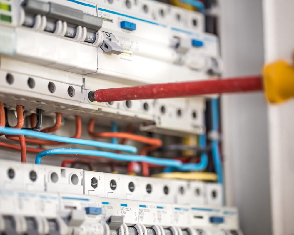 A close-up of a red screwdriver adjusting a circuit breaker in a home electrical panel with various colored wires.